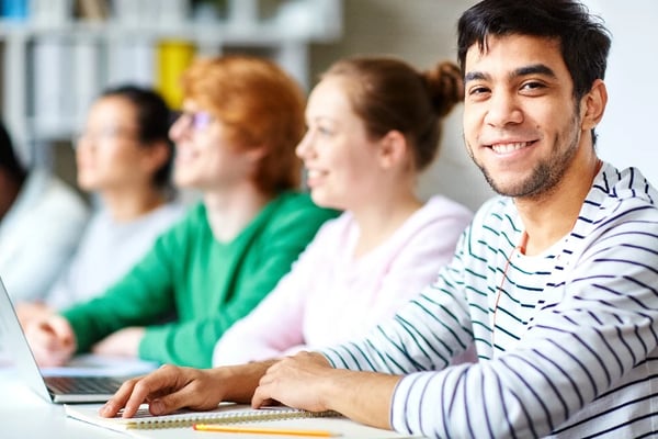 Four people writing notes in a classroom with one person smiling at the camera.