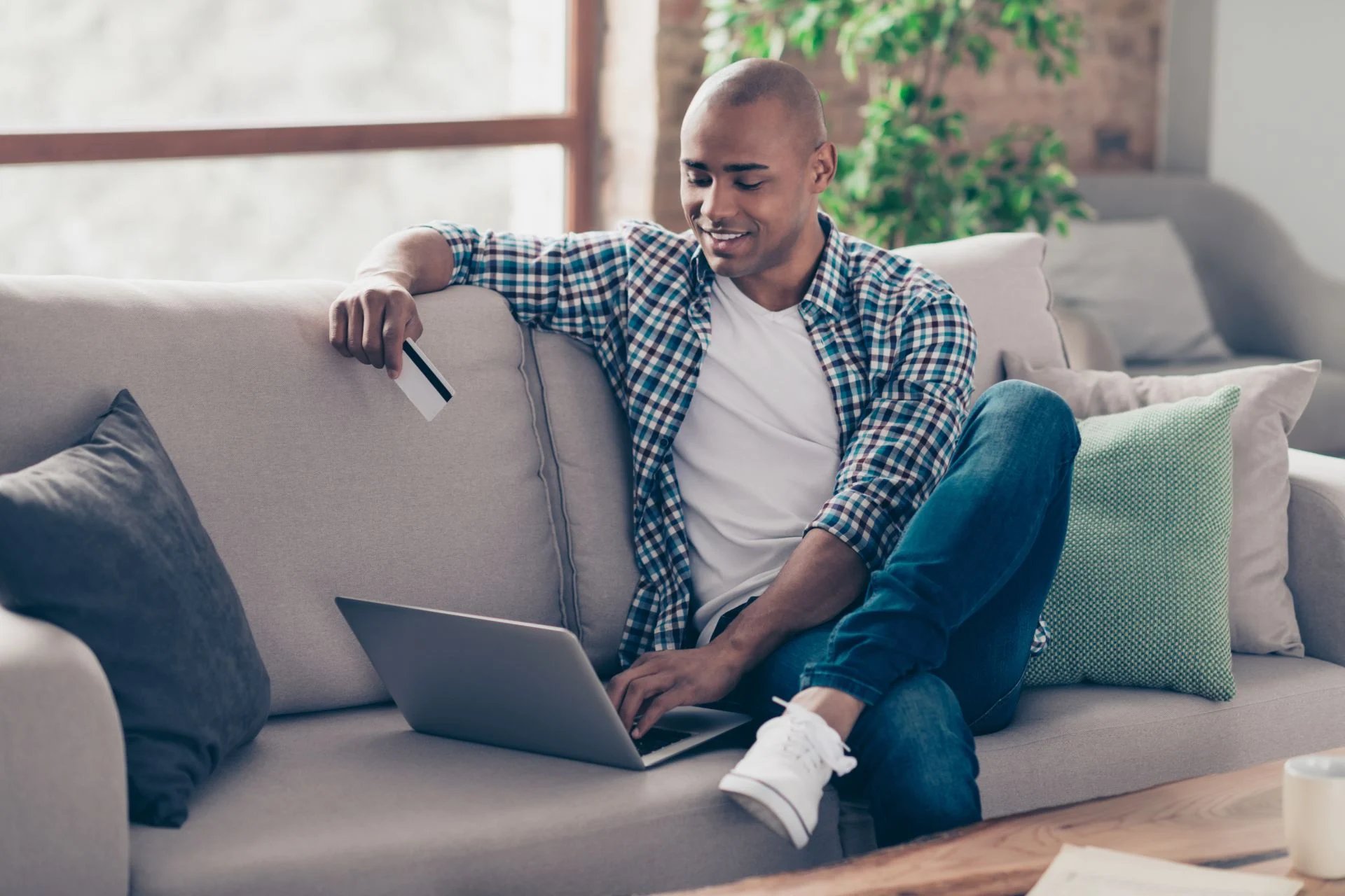 A man sitting on a couch with a credit card in hand about to enroll in a course through the student course registration software on his computer. 